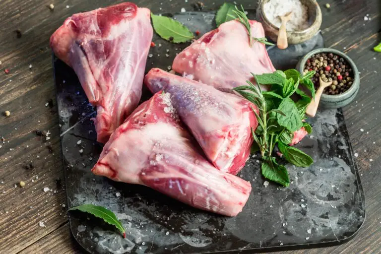 Four raw lamb shanks sprinkled with coarse salt are arranged on a dark chopping board, surrounded by bay leaves, fresh herbs, a bowl of salt, and a bowl of peppercorns on a rustic wooden surface.