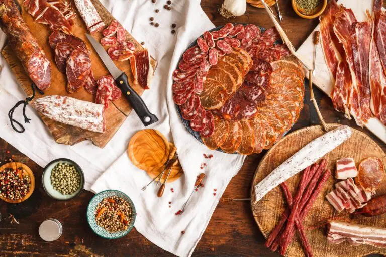A variety of cured meats, sausages, and salamis are arranged on wooden boards and plates, surrounded by spices, herbs, and kitchen utensils on a rustic wooden table.
