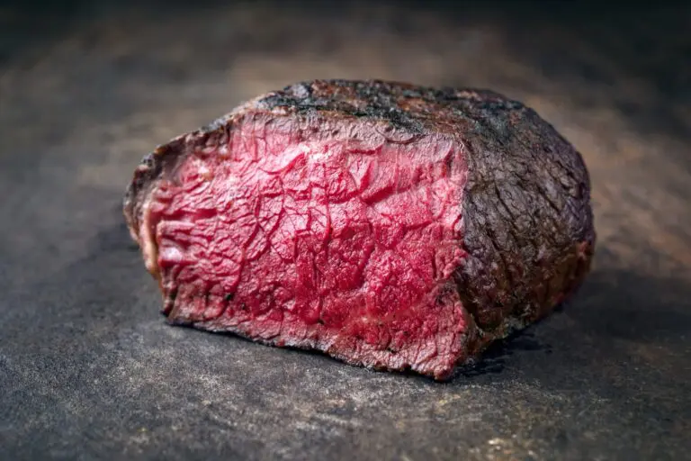 A close-up of a thick, juicy steak cooked rare, showing a charred brown exterior and a red, moist interior on a dark surface.
