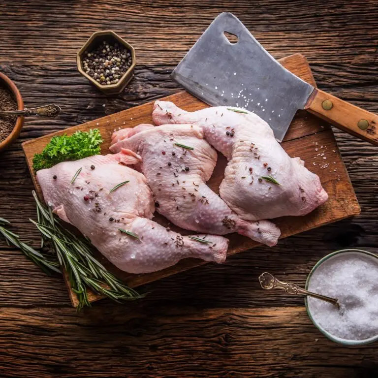 Raw chicken legs seasoned with peppercorns and rosemary on a wooden chopping board, surrounded by herbs, a bowl of peppercorns, a bowl of salt, and a large cleaver, all on a rustic wooden table.